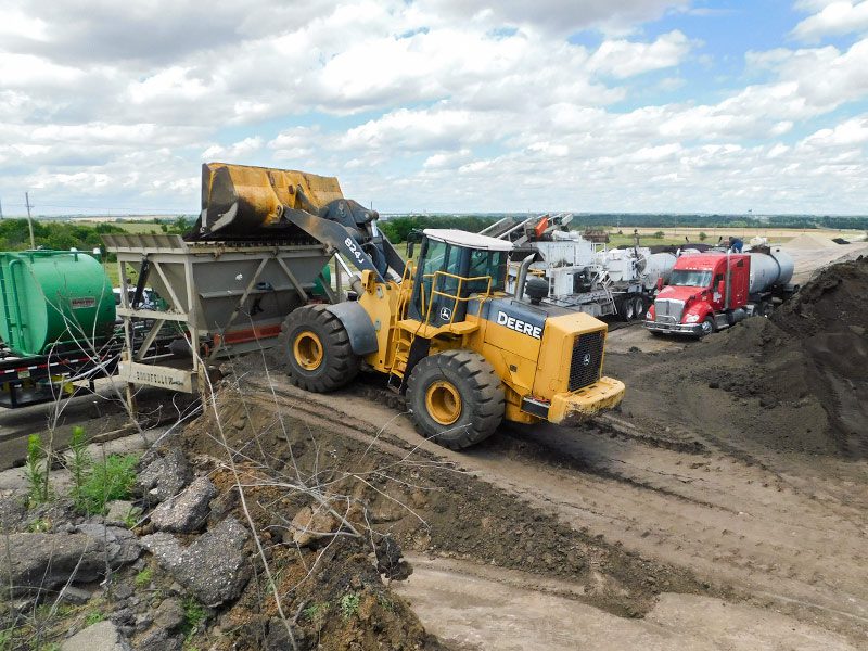 Tractor loading reclaimed asphalt pavement into a truck as part of the cold central plant recycling process.