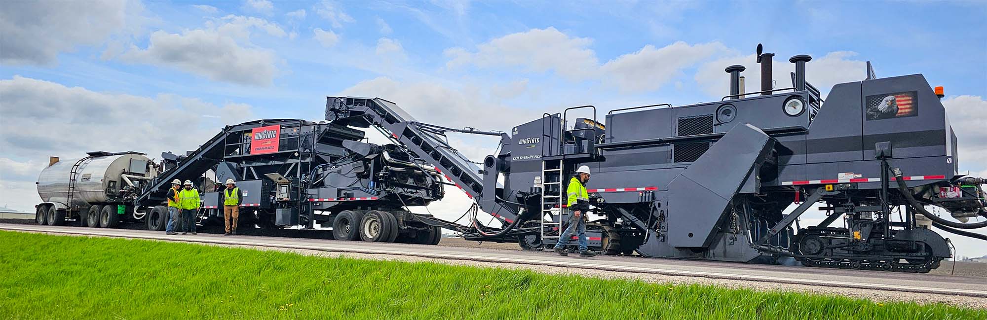 midstate-surfacecycle-cold-in-place-recycling-cir-banner Cold In-Place Recycling (CIR) train working on a road construction site, with grassy fields and cloudy skies.